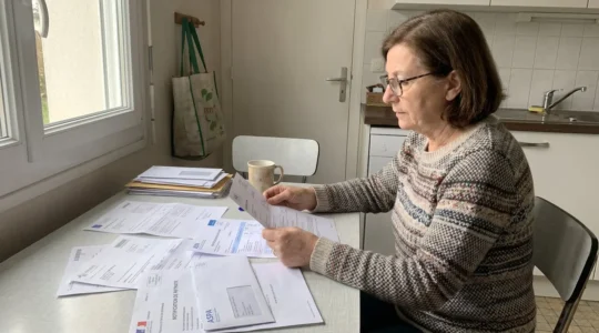 Une femme senior assise à une table de cuisine consulte des documents de retraite et des formulaires ASPA à la lumière naturelle